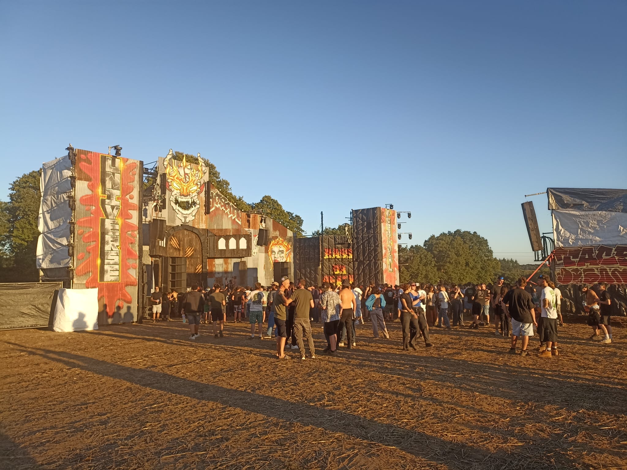 Festival techno en plein air au coucher du soleil, scène décorée en tôle graffée et public devant le mur de son