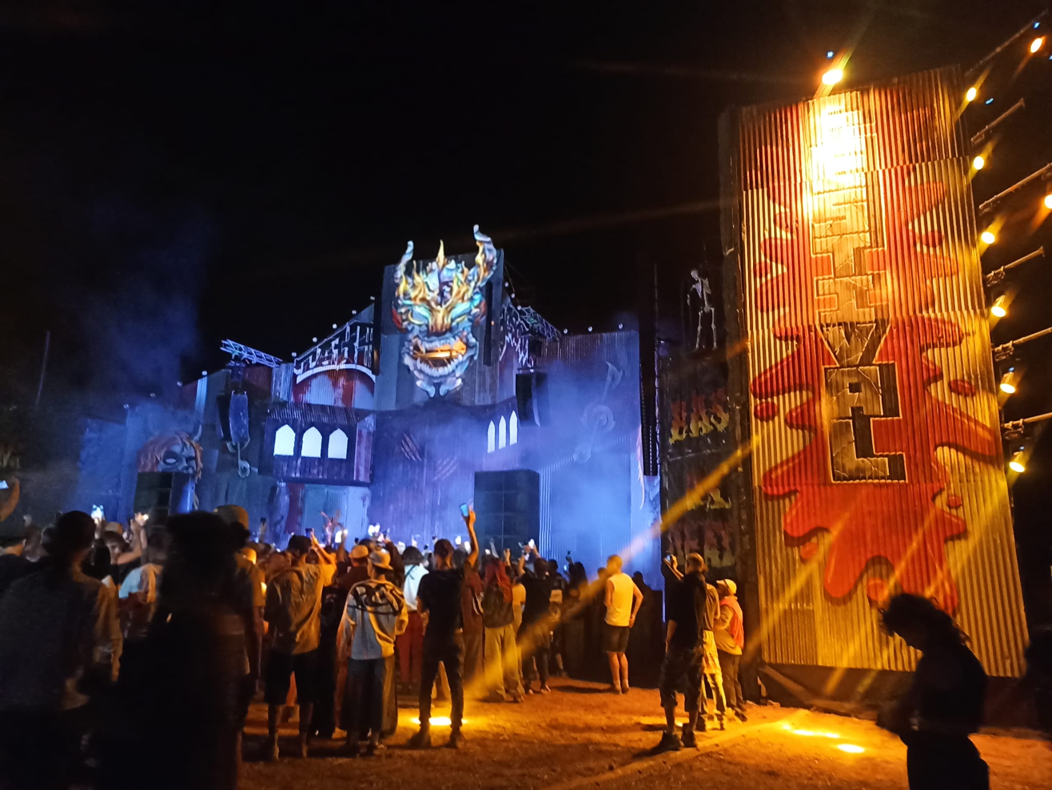 Scène de festival techno de nuit avec décor de démon enflammé, fumée bleue et foule de teufeurs devant le sound system